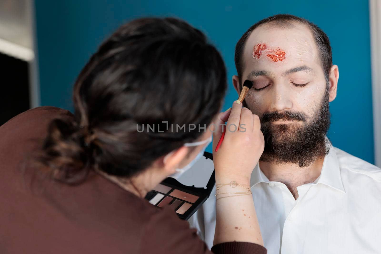Artist using makeup effects to create zombie costume and scary dramatic corpse look. Man looking deceased and infected after having bloody scars and monster face, creepy design.