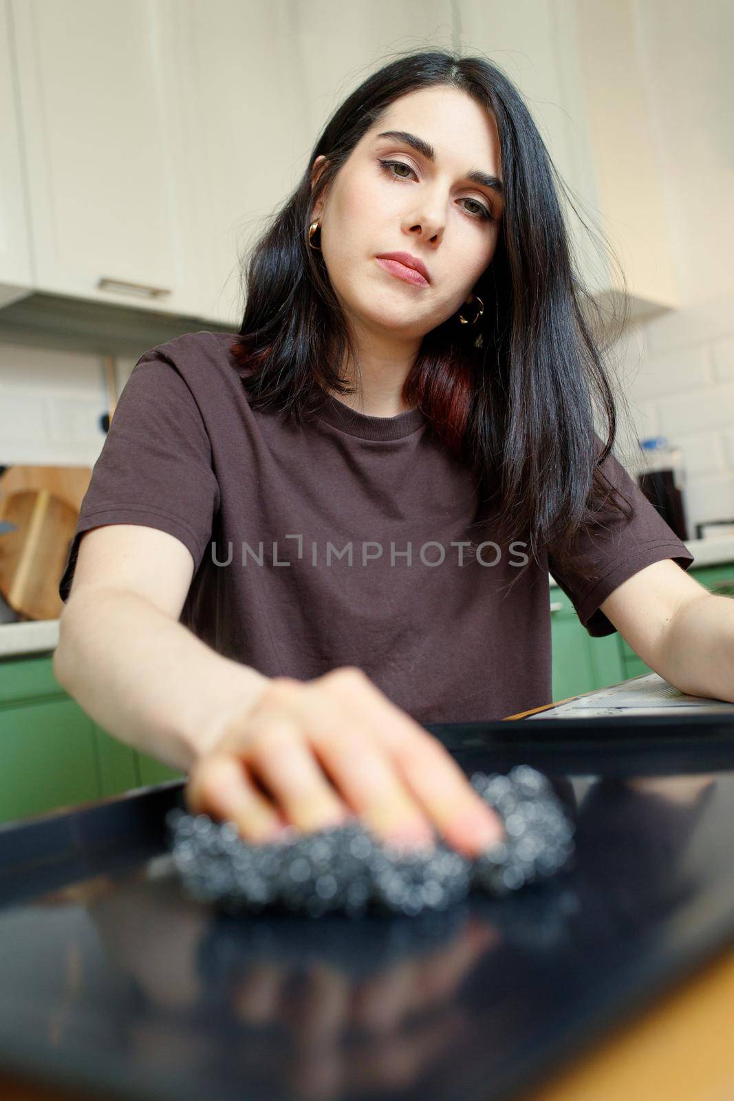 Tired young woman with a metal sponge for cleaning, washes a baking sheet in the kitchen, selective focus, vertical.