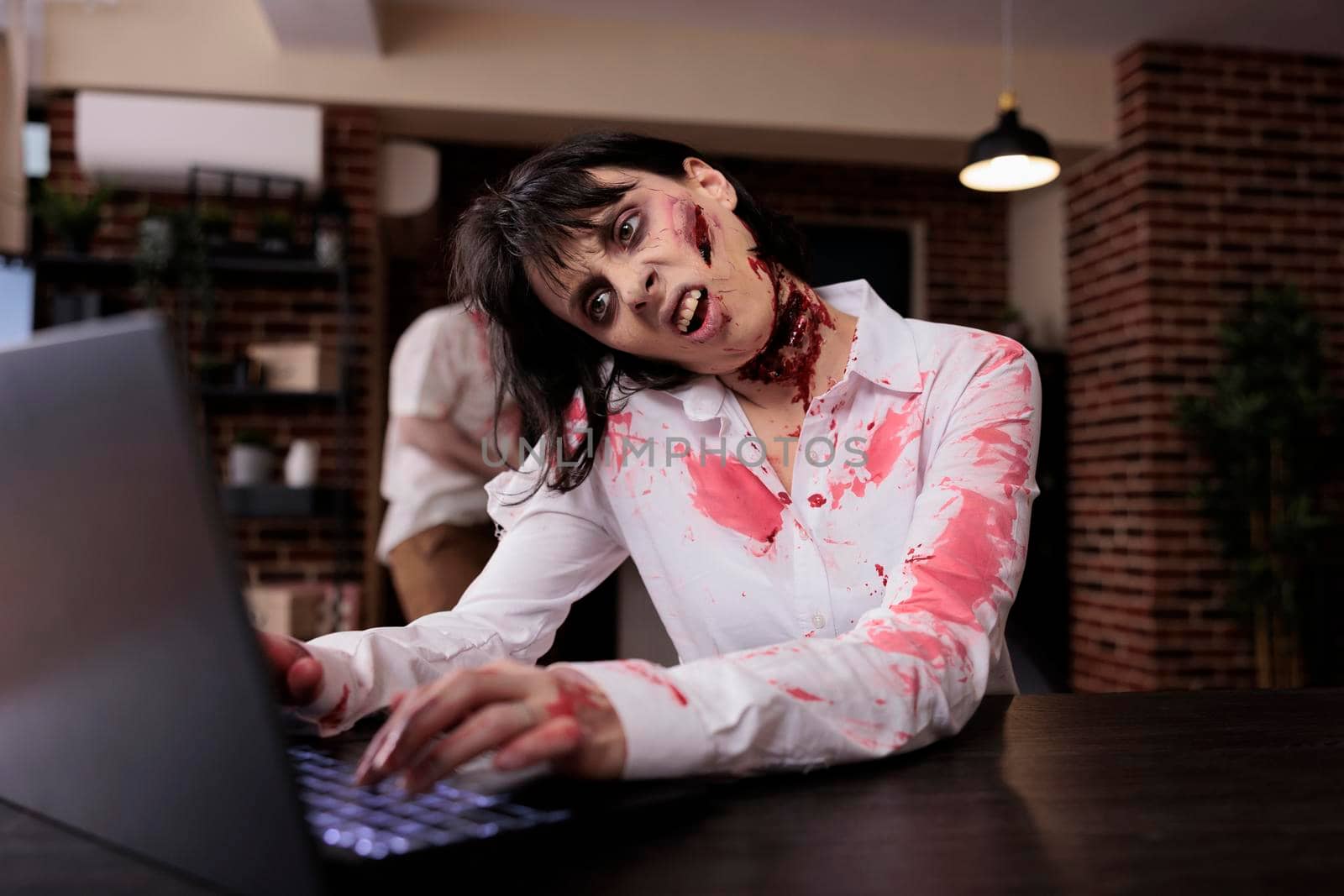 Aggressive devil corpse looking at laptop, sitting at office desk and being dangerous. Brain eating woman monster with bloody gory wounds being dramatic and eerie with mouth open.
