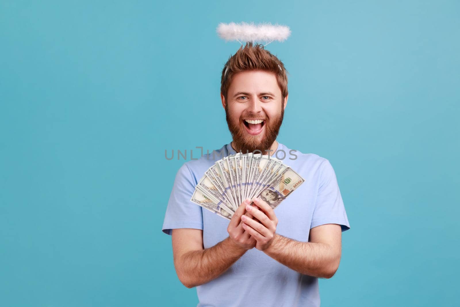 Portrait of excited bearded angelic man with holy nimbus over head holding fan of dollar bills and smiling happily, happy to have money. Indoor studio shot isolated on blue background.