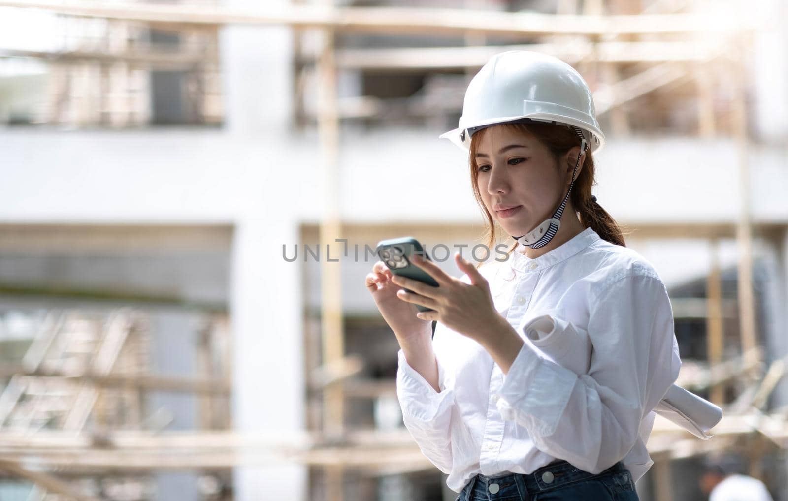 Architect with a blueprints at a construction site. Portrait of woman constructor wearing white helmet and safety yellow vest. Upset sad, skeptical, serious woman looking at the phone screen outdoors. by wichayada