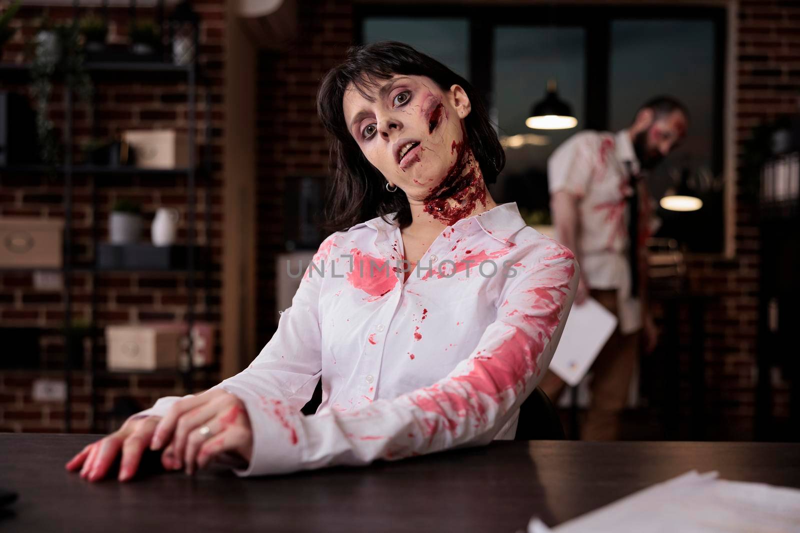 Portrait of evil zombie with wounds in office, sitting at desk to work on laptop being terrifying and spooky. Cruel possessed brain eating monster with bloody mouth open, walking dead devil.
