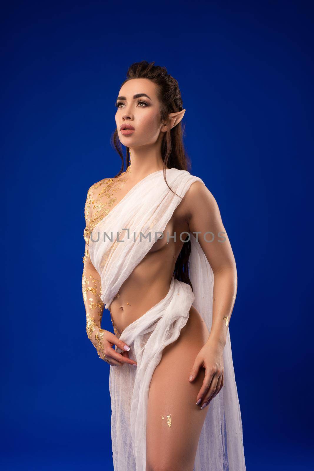 Gorgeous young female model with long dark hair and elf ears in white dress looking away pensively against blue background