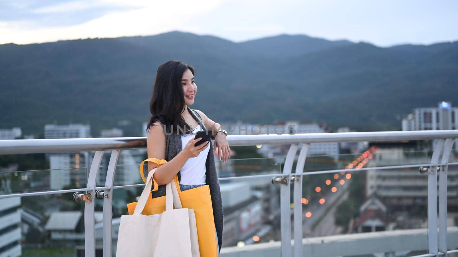 Trendy young asian woman with with shopping bags standing on terrace against background of cityscape at sunset by prathanchorruangsak