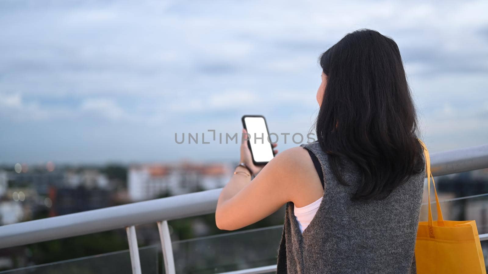 Back view of young woman using mobile phone while standing at terrace with beautiful evening sky in background.