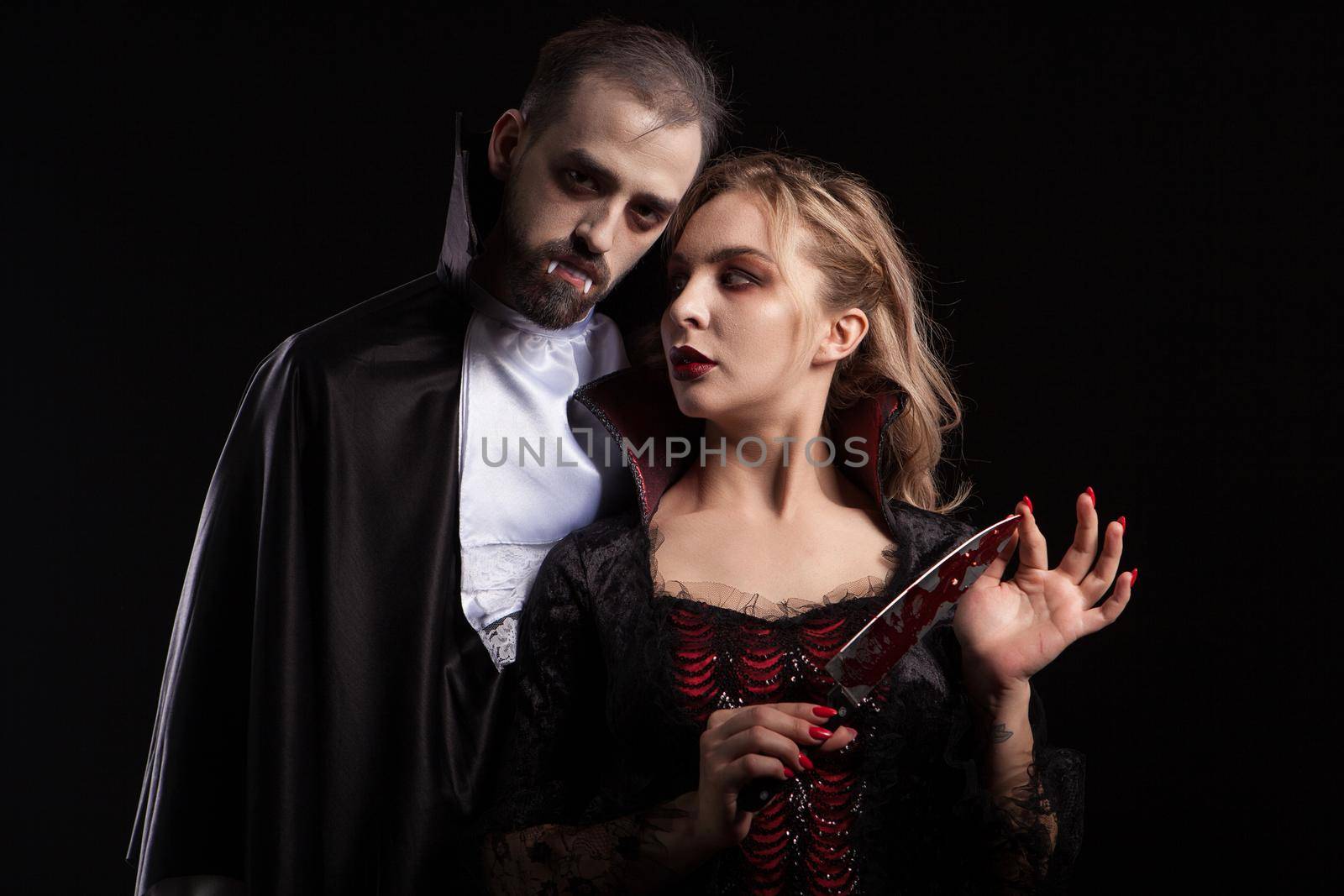 Beautiful young vampire woman with a blade covered in blood looking at her man dressed up like Dracula for halloween by DCStudio
