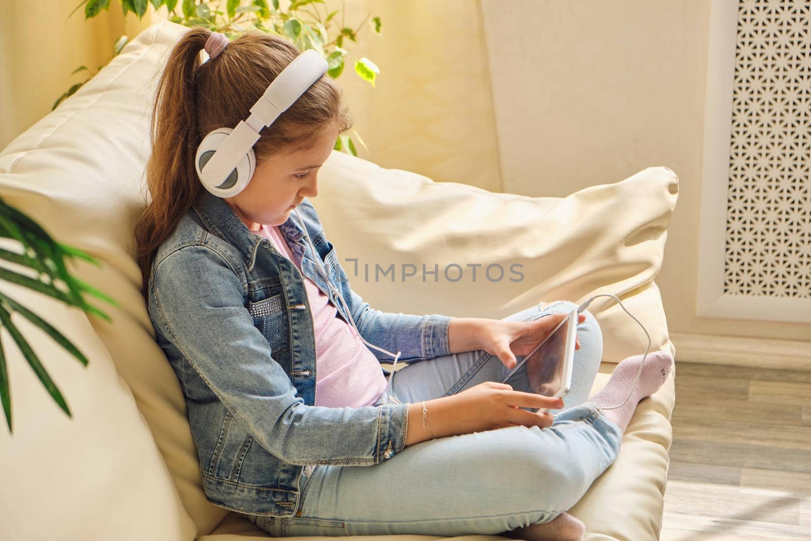 Cute little girl in casual clothes and headphones using a tablet and listening to music while sitting on a sofa in the room.