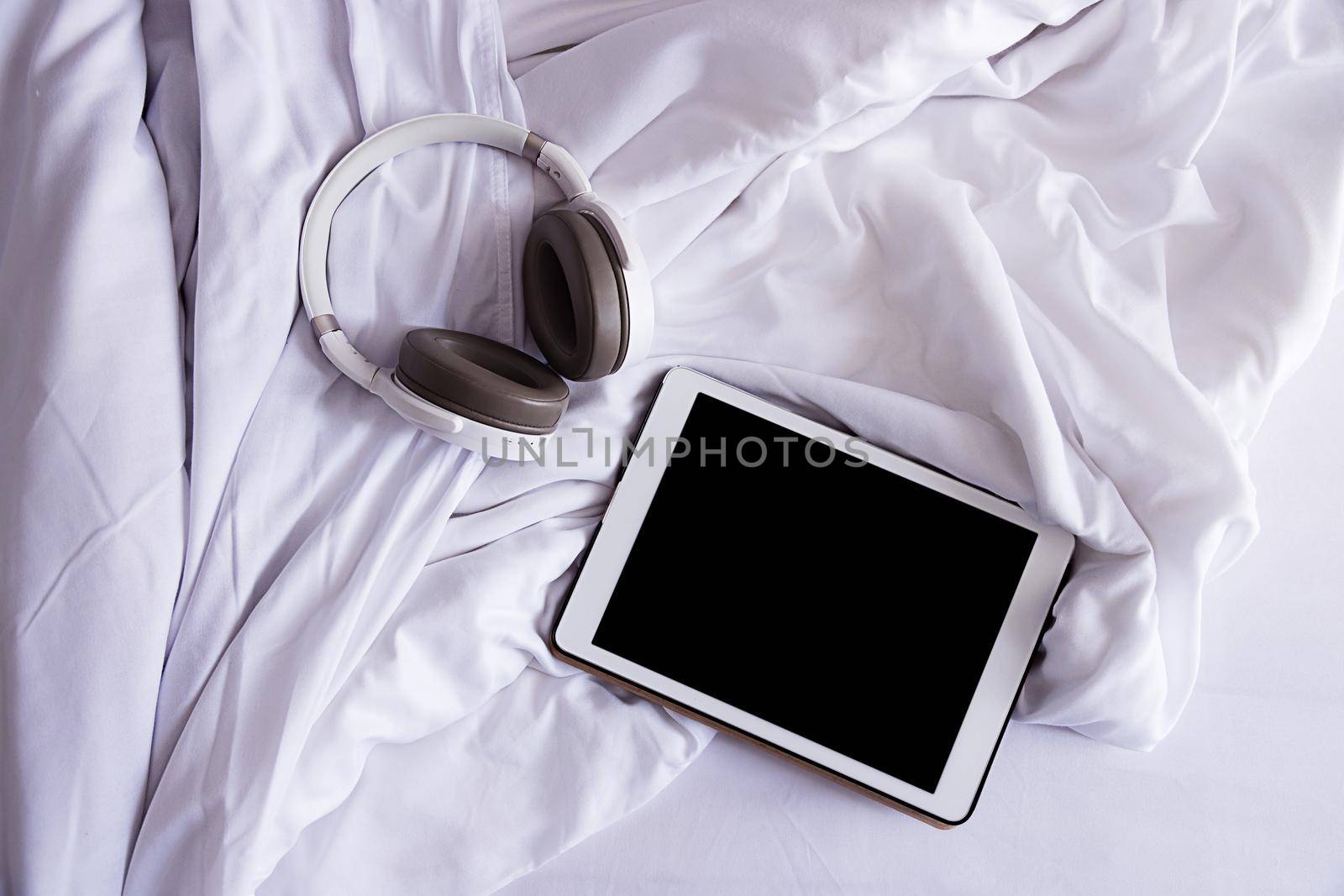 a tablet and headphones lie on a crumpled white bed, top view, close-up by Ramanouskaya