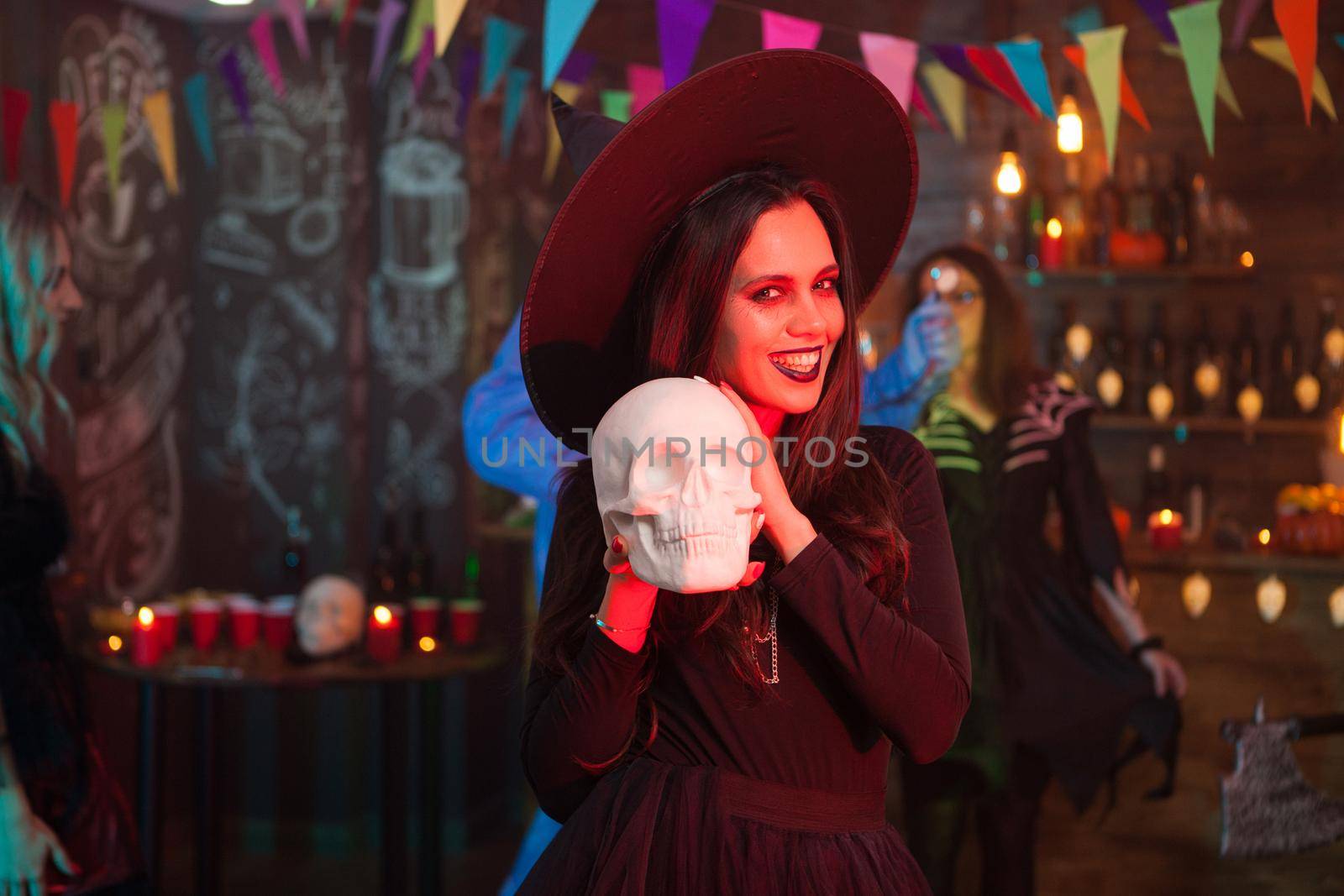 Attractive woman dressed like a witch for halloween smiling with a human skull by DCStudio
