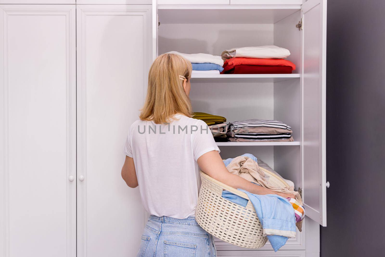 woman stands in front of closet with clothes, collects dirty things for washing by Ramanouskaya
