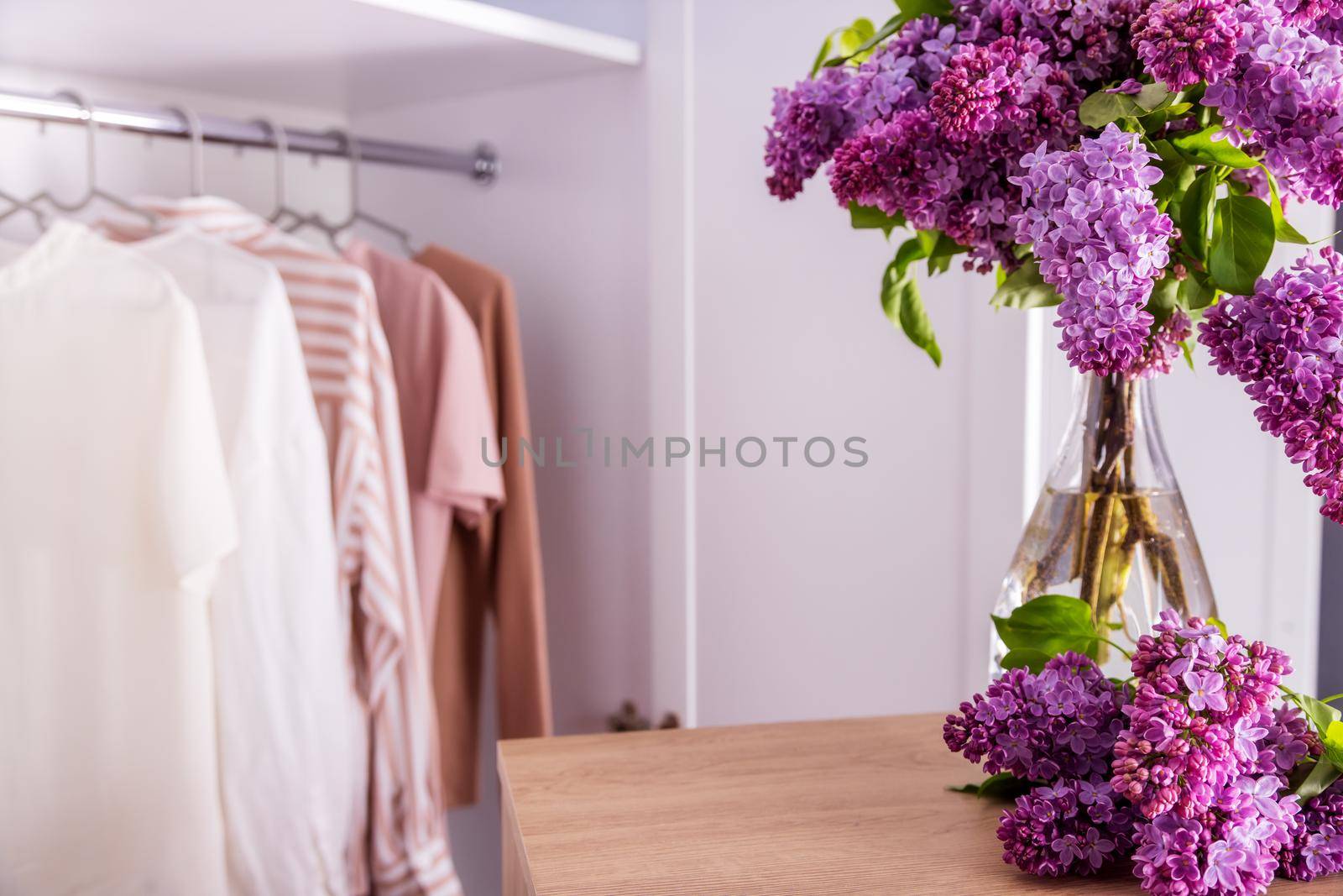 Women's clothes in pink and beige on a white hanger in an open closet. Wooden table in the foreground with copy paste for text or product. on a wooden table there is a vase with lilac branches