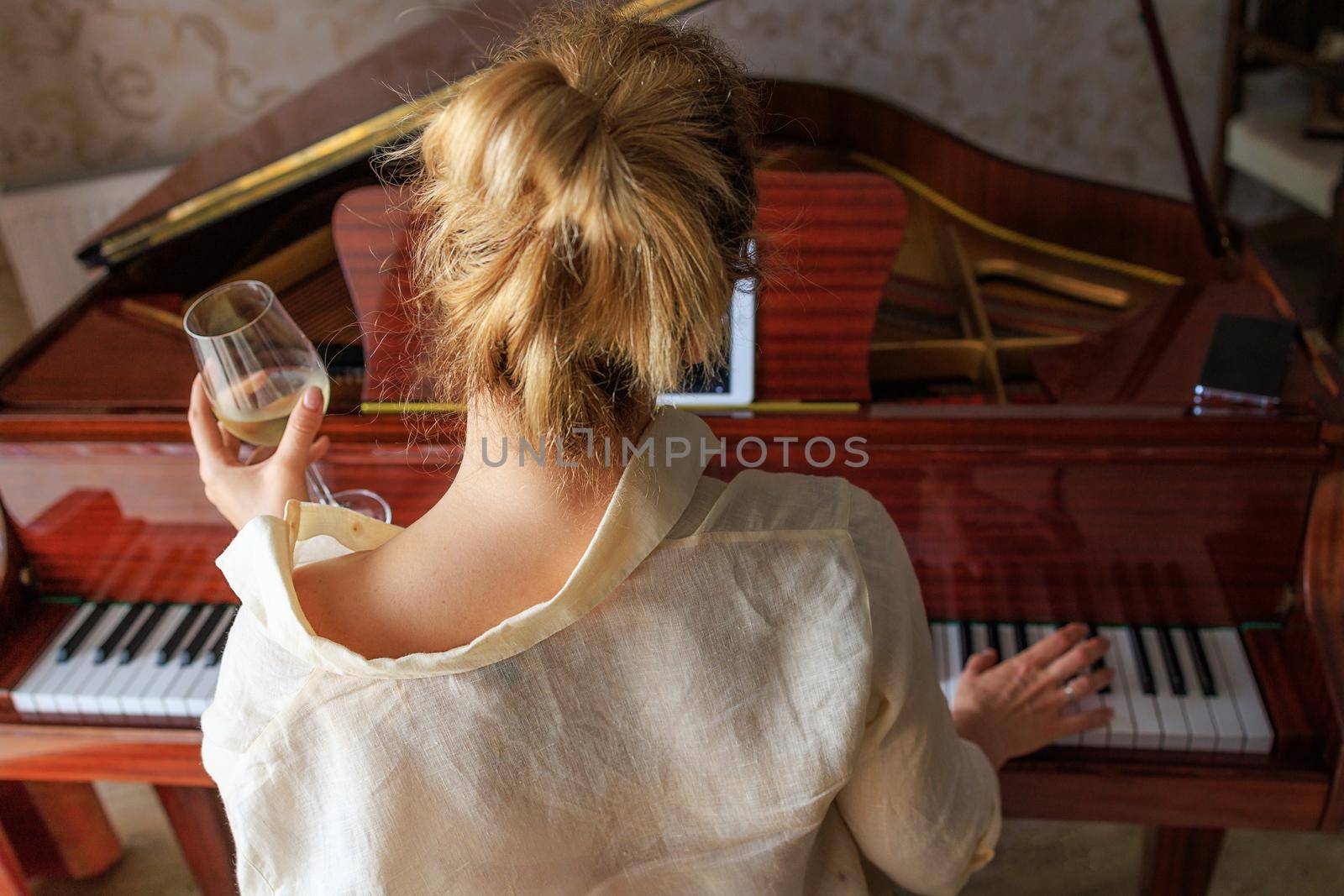 sad young blonde plays music on a retro piano, looks at notes in tablet. Sits back. The girl is holding a glass of wine in her hands. The concept of loneliness, depression, alcoholism in young people