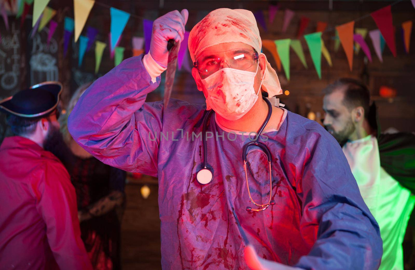 Portrait of spooky doctor with a knife on his hand looking into the camera at a halloween celebration. Man in dracula costume in the background.