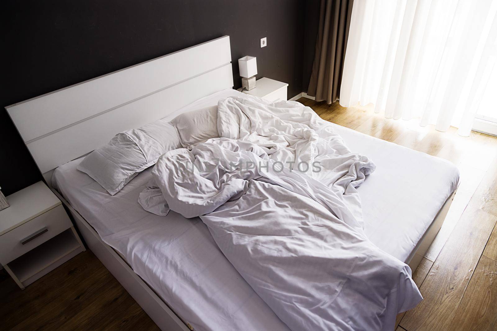 Crumpled white bed with set of white bed linen in bedroom in the early morning by Ramanouskaya