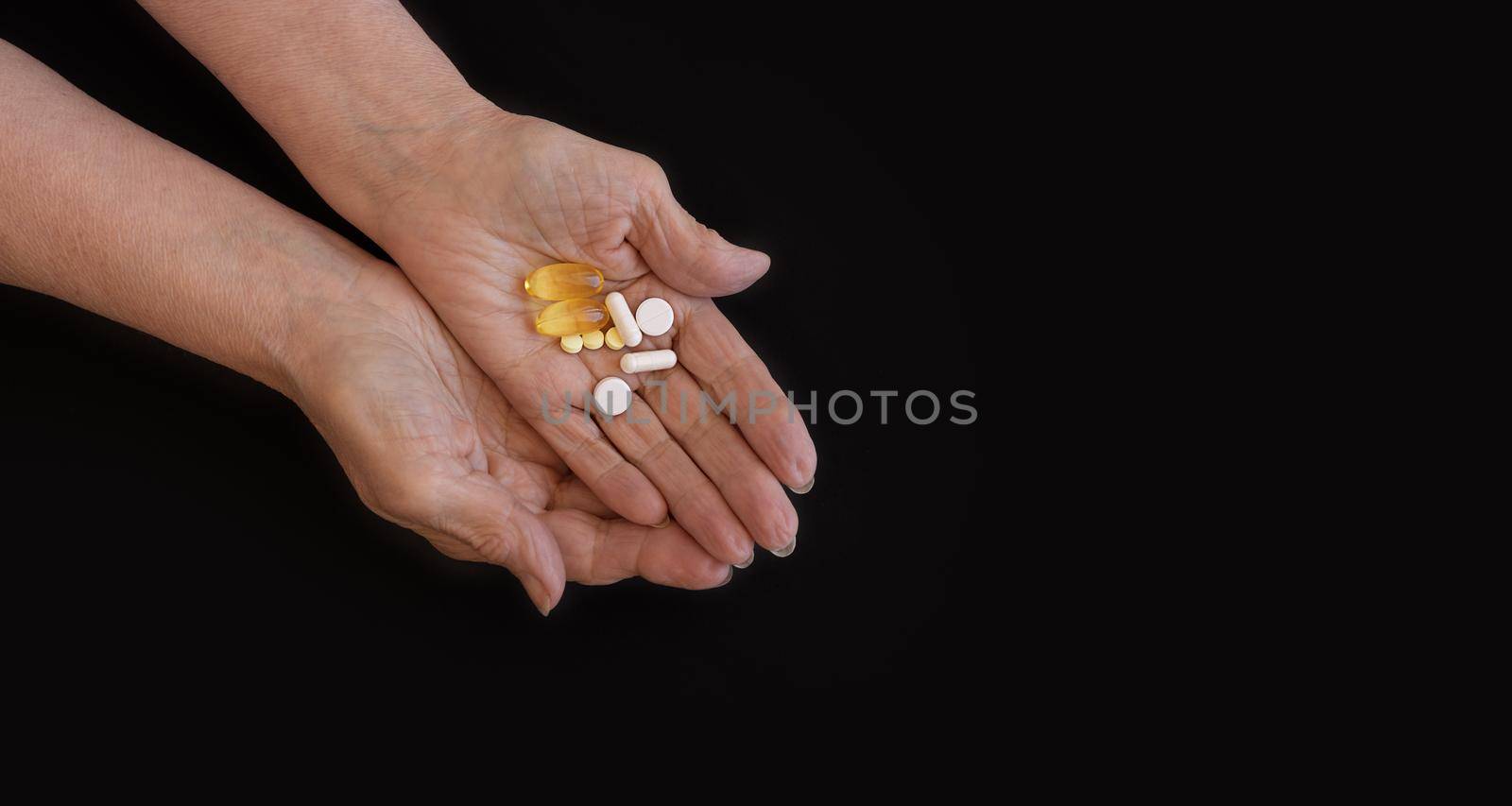 Close-up of elderly woman's hand holding a handful of different pills. Black background, copy paste for your text. The concept of health prevention, taking vitamins and minerals, maintaining the body