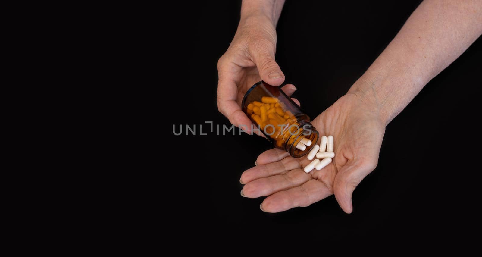The hands of an elderly woman pour a pill from a jar into her palm. Black background, copy paste for your text. The concept of medicine, diseases of the elderly, prevention, taking vitamins