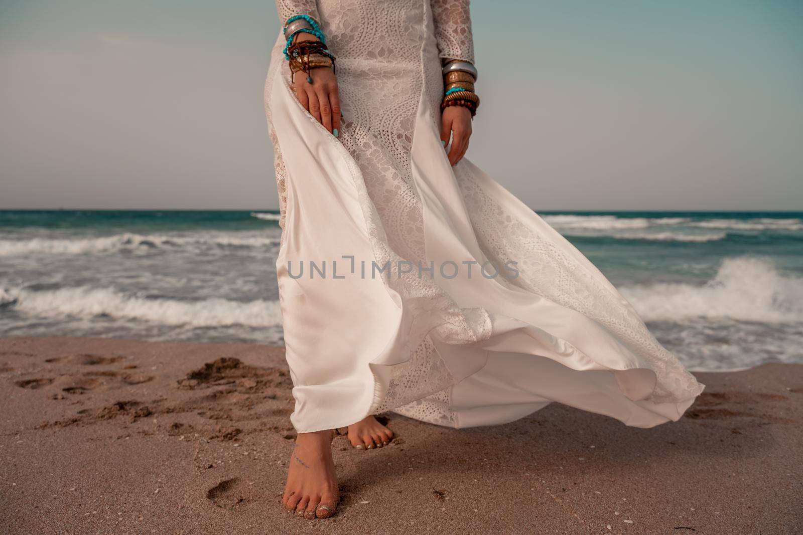 Model in boho style in a white long dress and silver jewelry on the beach. Her hair is braided, and there are many bracelets on her arms