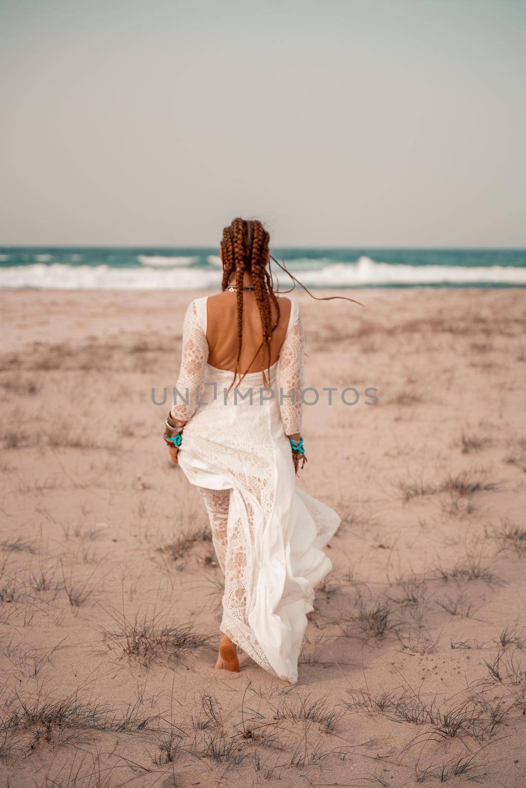 Model in boho style in a white long dress and silver jewelry on the beach. Her hair is braided, and there are many bracelets on her arms. by Matiunina