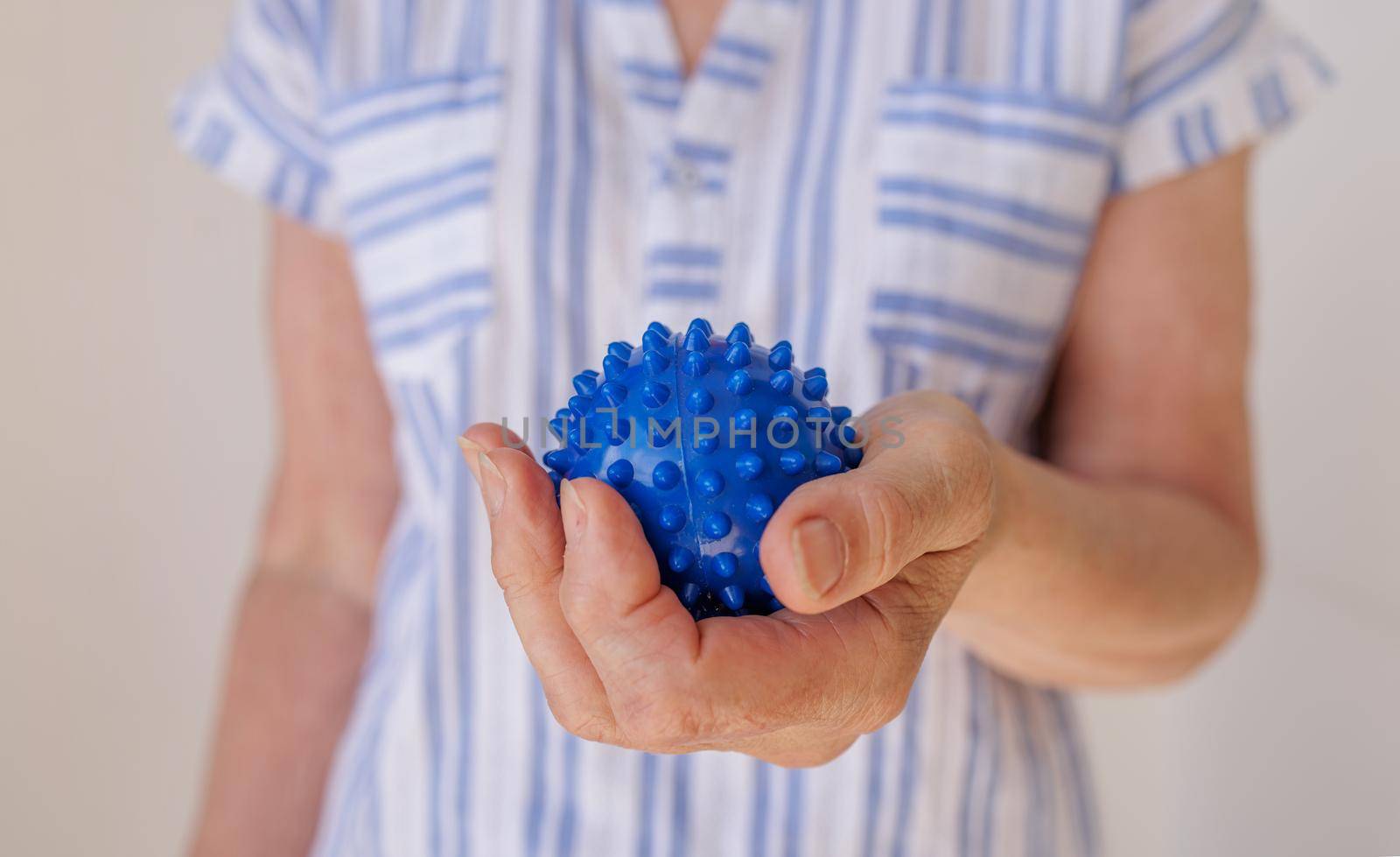 An elderly white woman in a blue T-shirt self-massages her hands with a prickly ball. Technique for helping your body with arthritis, parkinson, padagra and other joint diseases