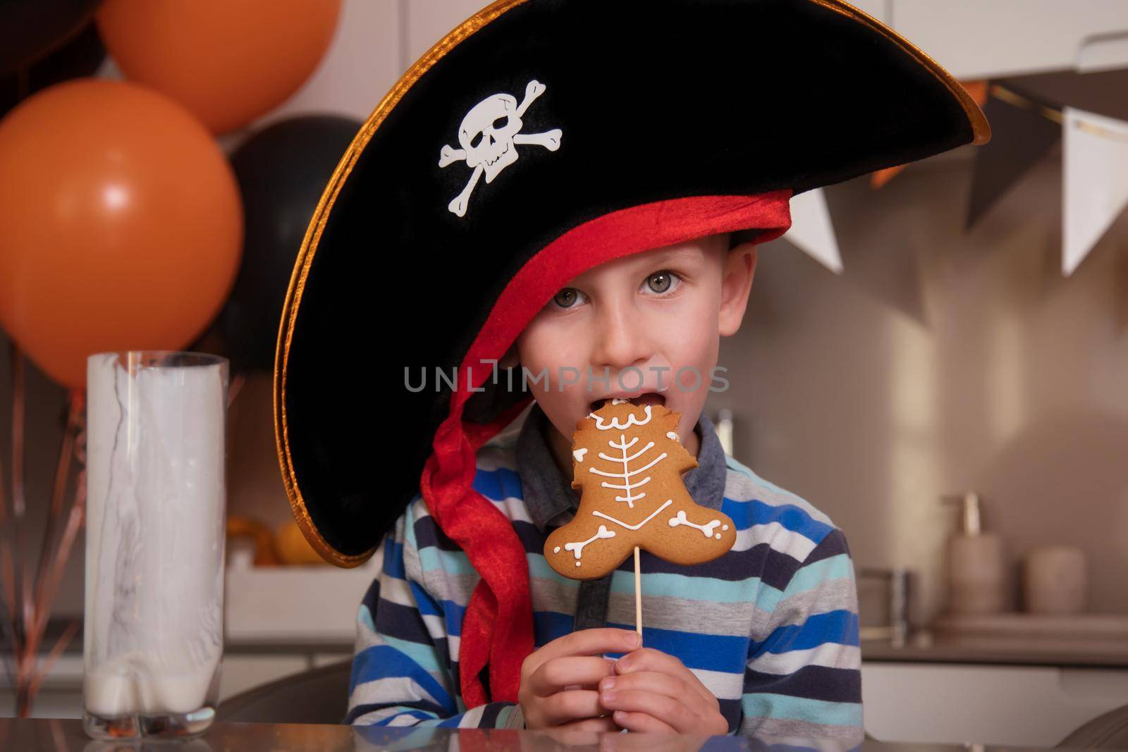 A boy dressed as a pirate eats gingerbread as a Halloween skeleton, drinks milk by Ramanouskaya