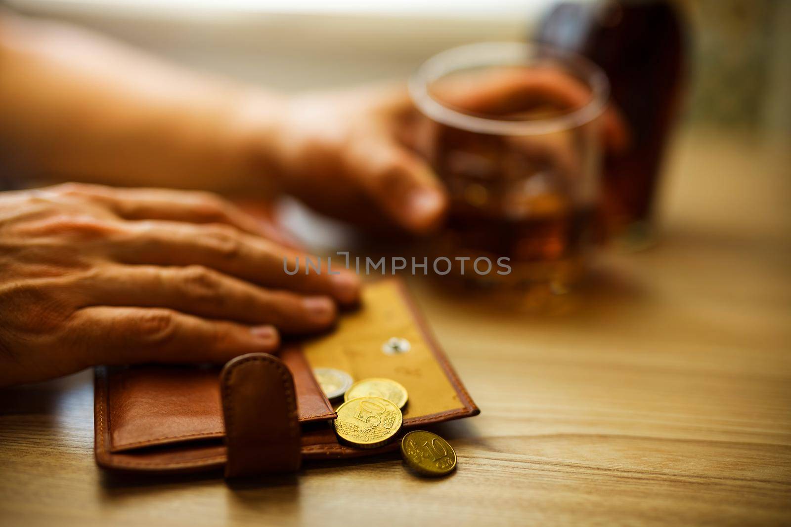 Fired man countsing his last money to drink expensive alcohol. Wrinkled alcoholic sits at brown wooden table with a glass of cognac with ice, on the table, empty wallet with coins