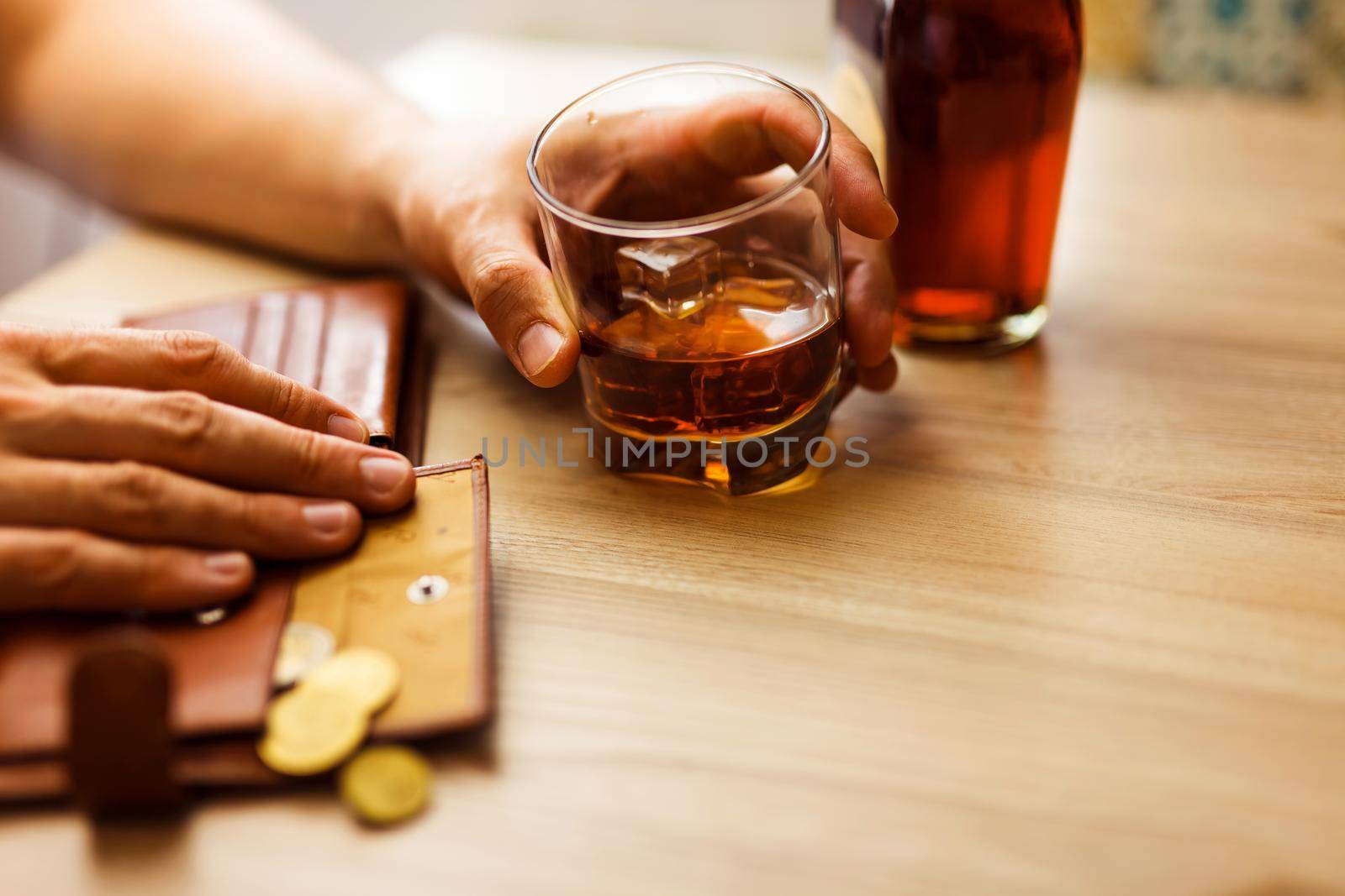 Fired man countsing his last money to drink expensive alcohol. Wrinkled alcoholic sits at brown wooden table with a glass of cognac with ice, on the table, empty wallet with coins