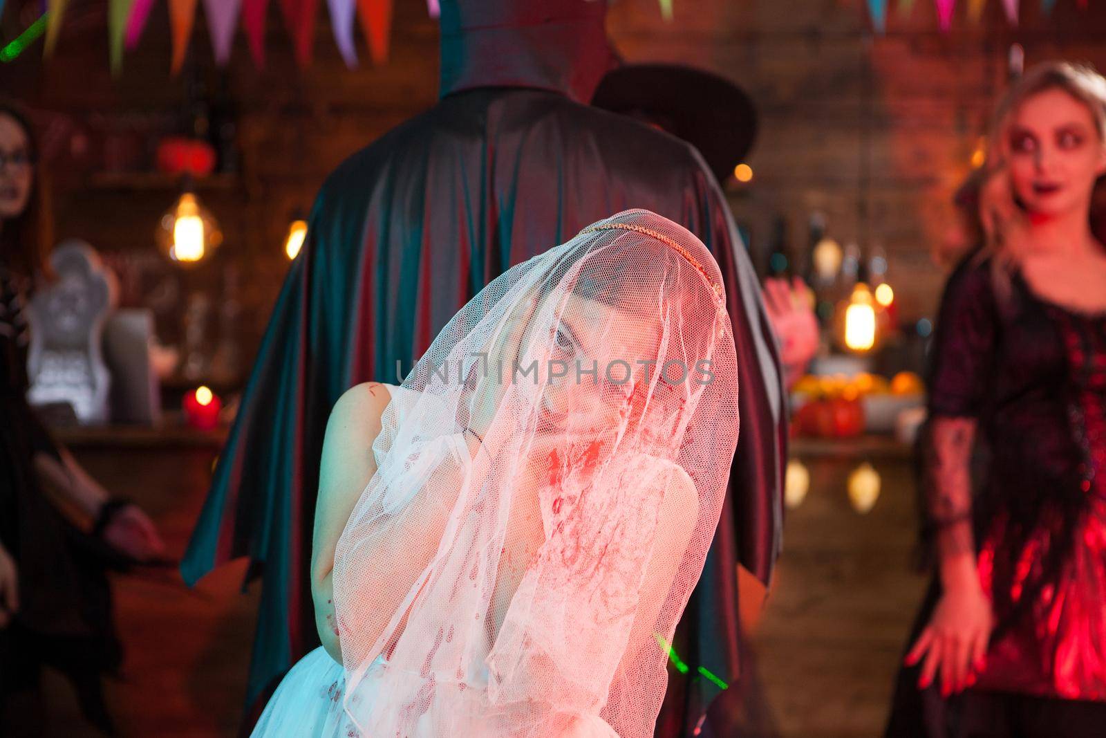 Little girl with no emotions dressed up like a bride at a halloween party. Celebrating Halloween. Man in dracula costume blurred in the background.