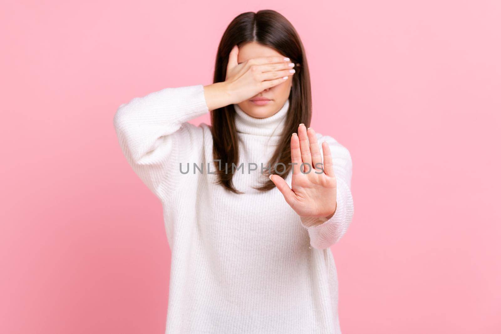 Confused woman covers eyes, raising hand to stop, feeling stressed, refusing to watch scary content, wearing white casual style sweater. Indoor studio shot isolated on pink background.