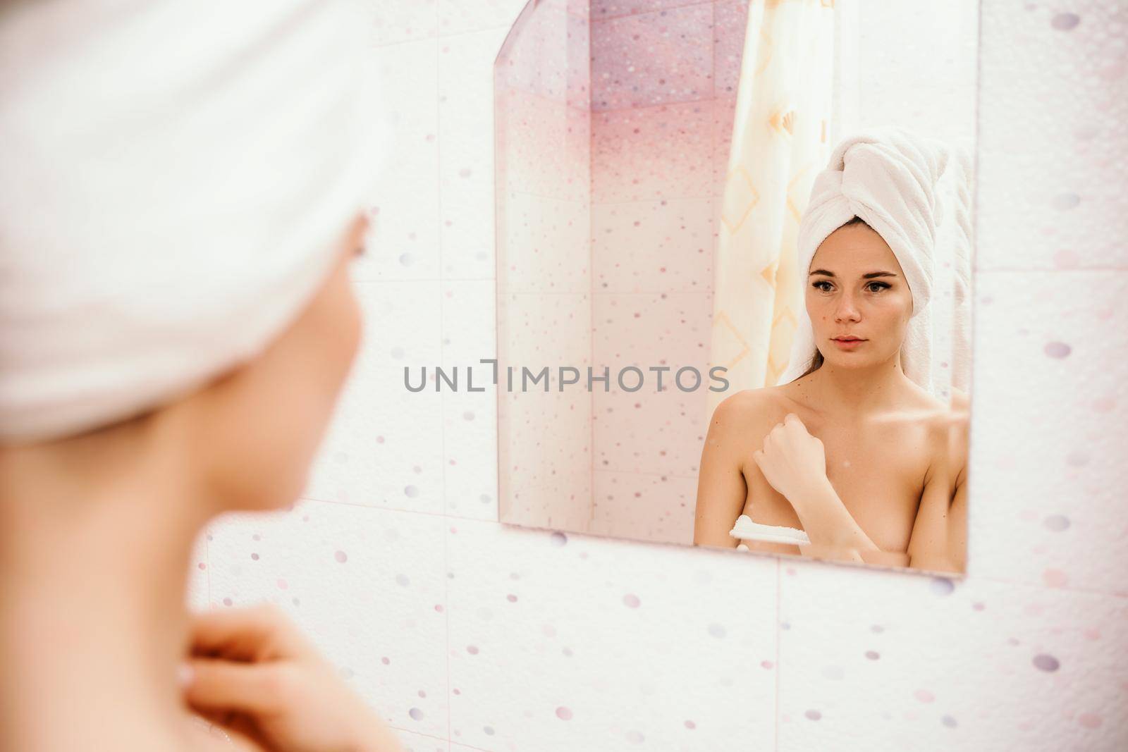 Portrait of young girl with towel on head in white bathroom looks and touches her face in the mirror and enjoys youth and hydration. Natural beauty, home care for problem skin.