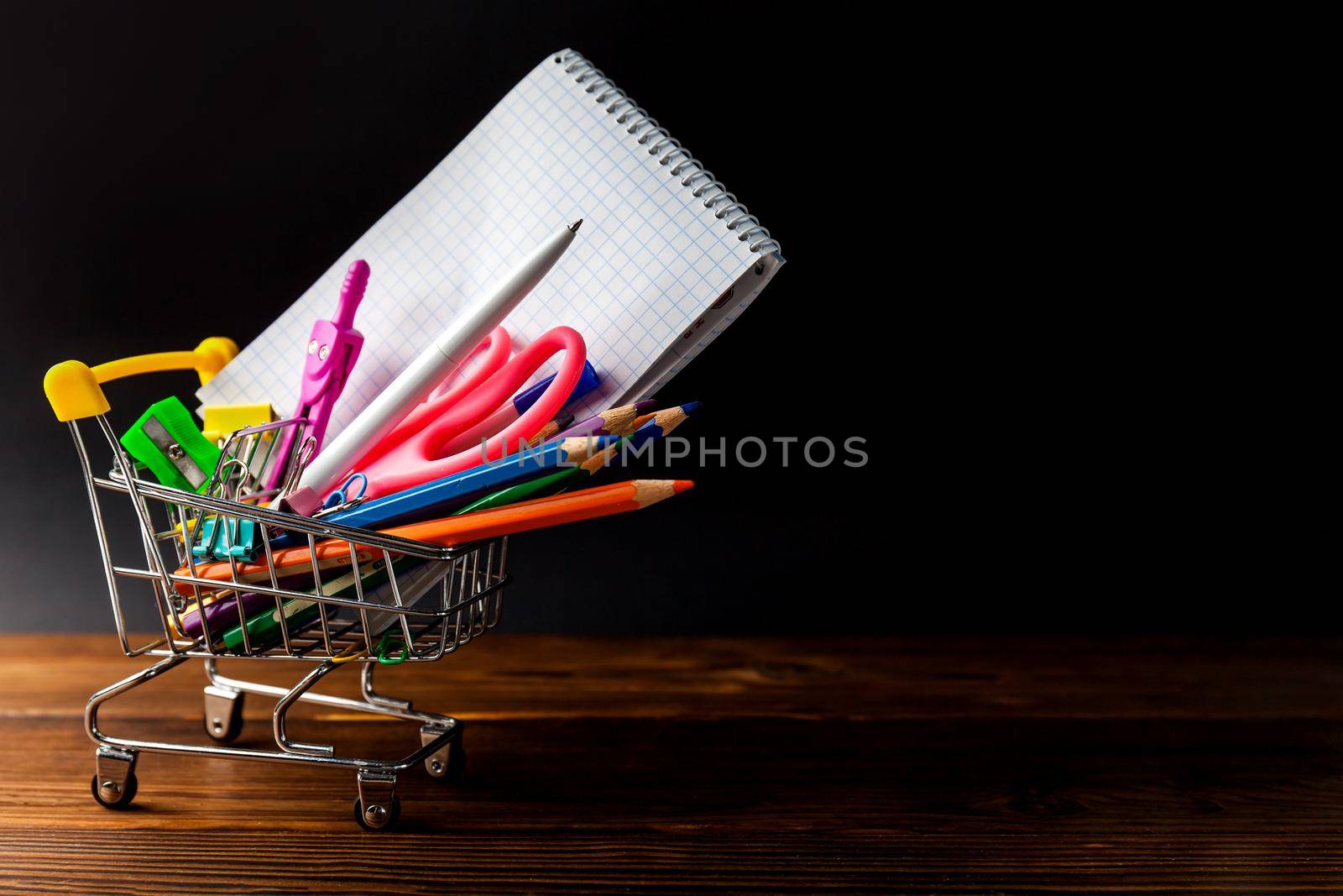Shopping cart full of stationery or office supplies on dark background. by InnaVlasova