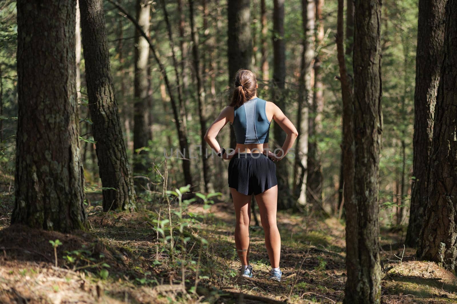 Rear view of female trail runner dressed in sportswear standing on trail in the forest and preparing to run. by apavlin