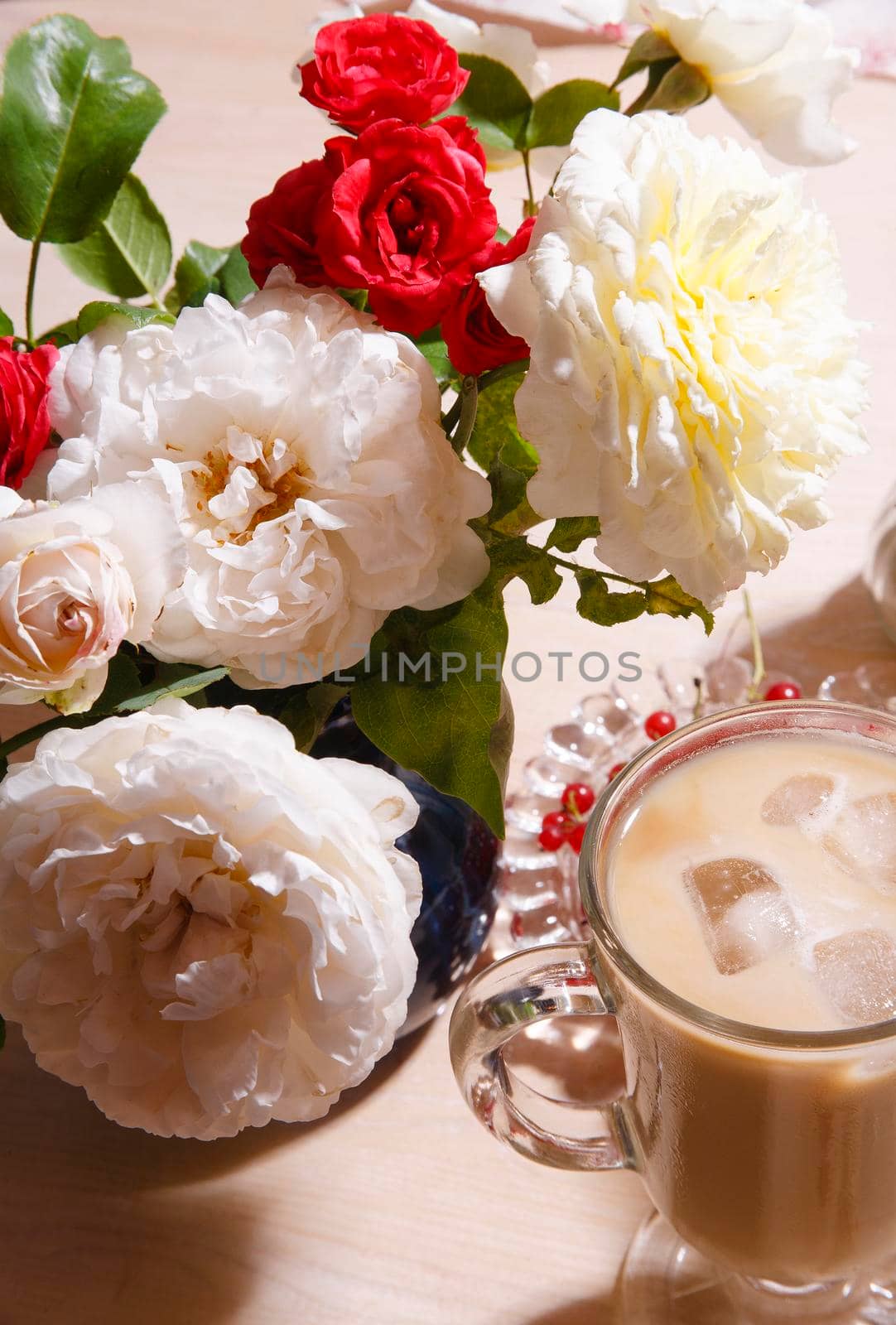 A cup of iced coffee with a bouquet of white, yellow and red roses on white table in the early summer morning, selective focus, hard light close up.