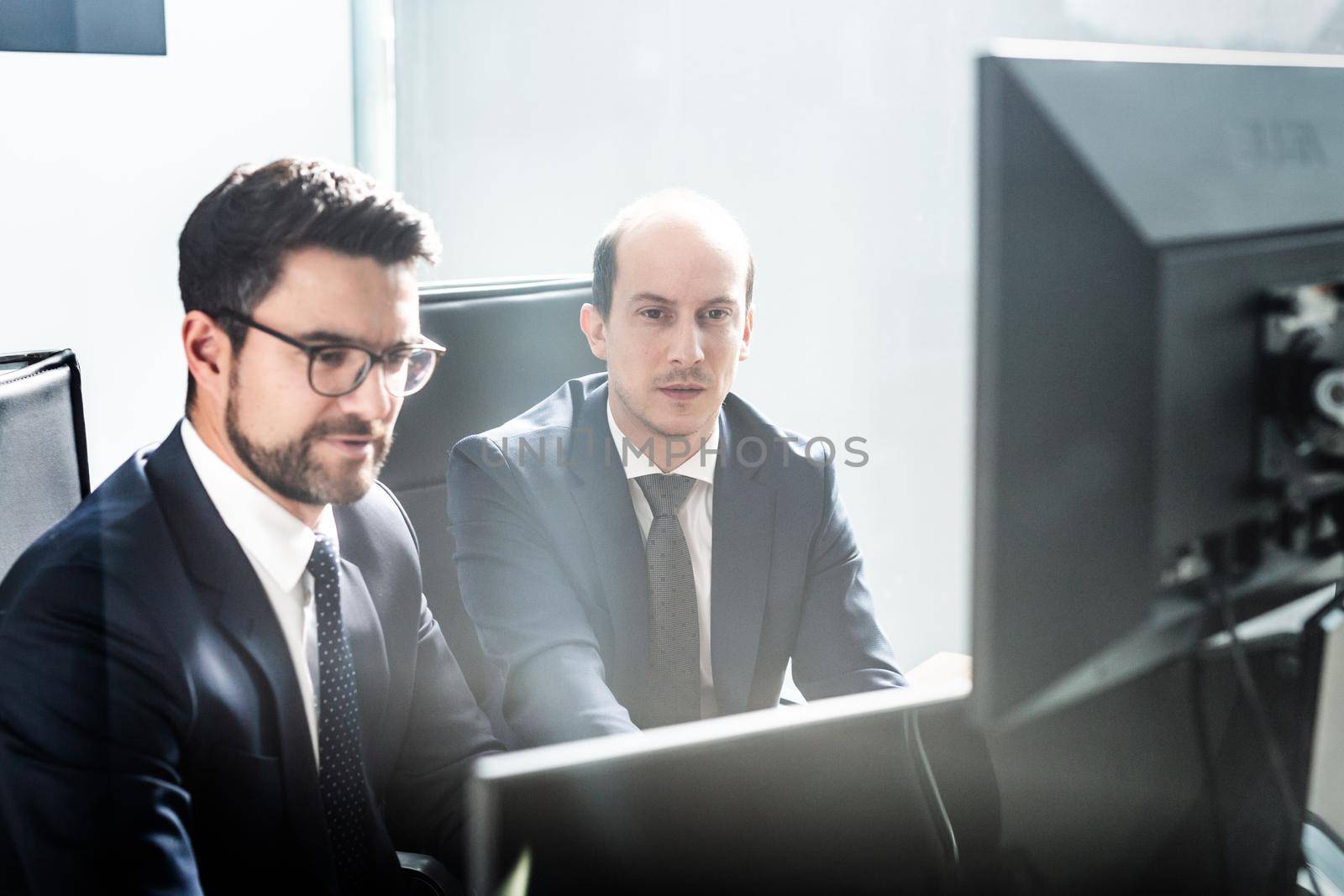 Image of two thoughtful businessmen looking at data on multiple computer screens, solving business issue at business meeting in modern corporate office. Business success concept.