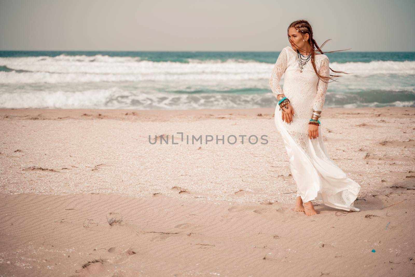 Model in boho style in a white long dress and silver jewelry on the beach. Her hair is braided, and there are many bracelets on her arms