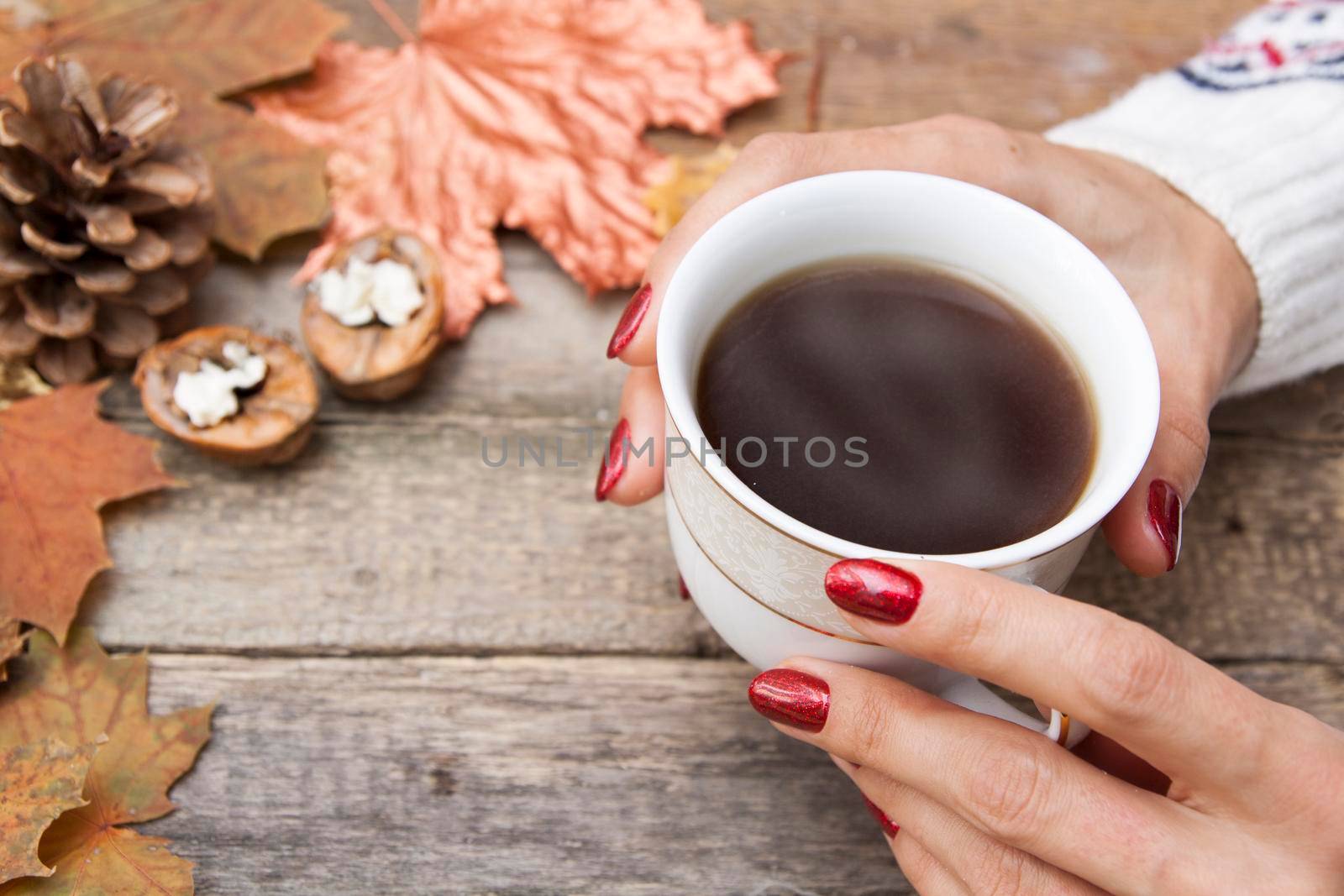 Female hands in a sweater warm their hands on a cup of hot coffee. On a wooden table are autumn leaves, nuts, cone.
