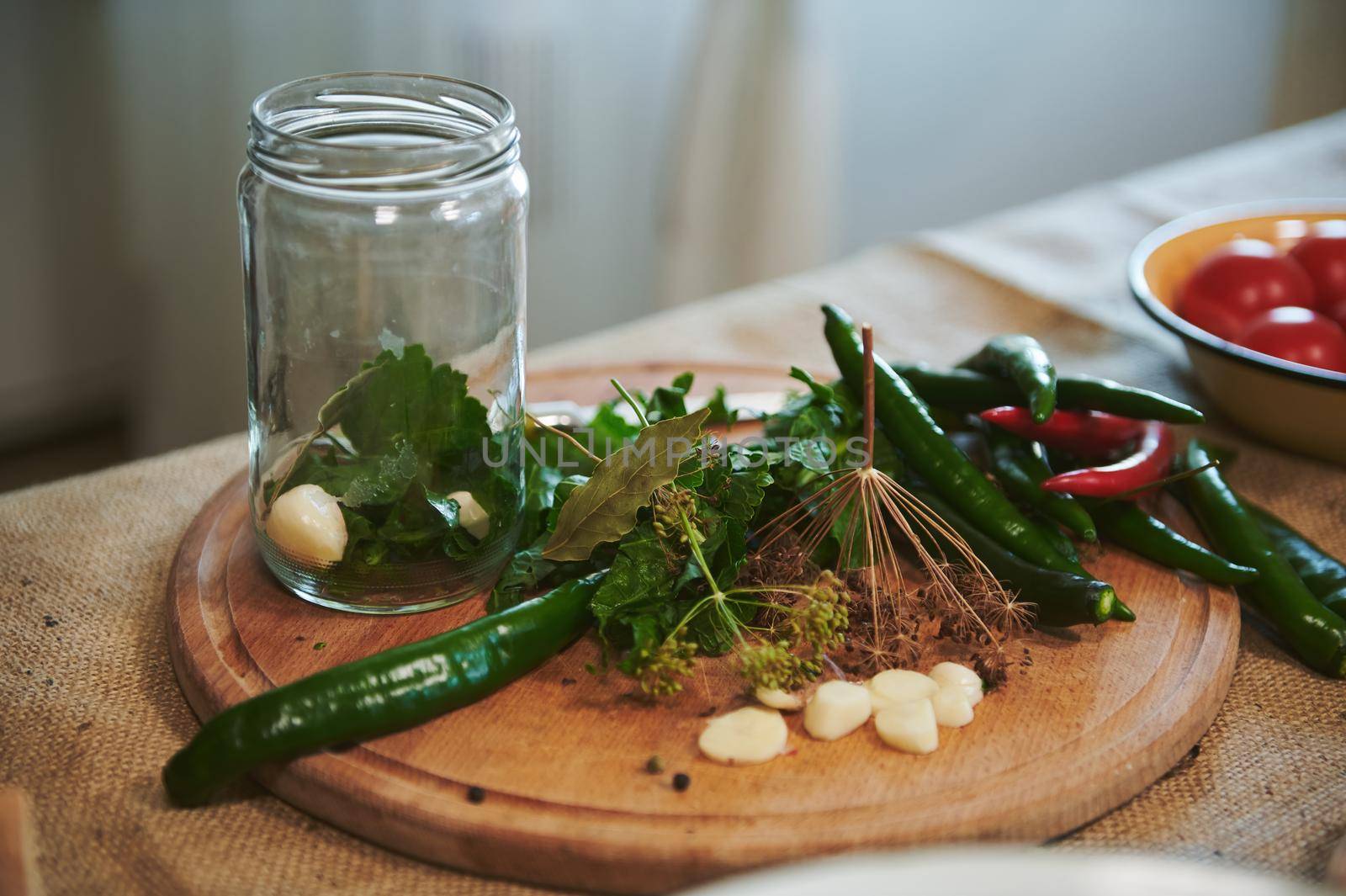 Still life with sterilized glass can for canning, peeled garlic cloves, chopped fresh culinary herbs and various chili peppers on a wooden chopping board, ready for pickling in the home kitchen