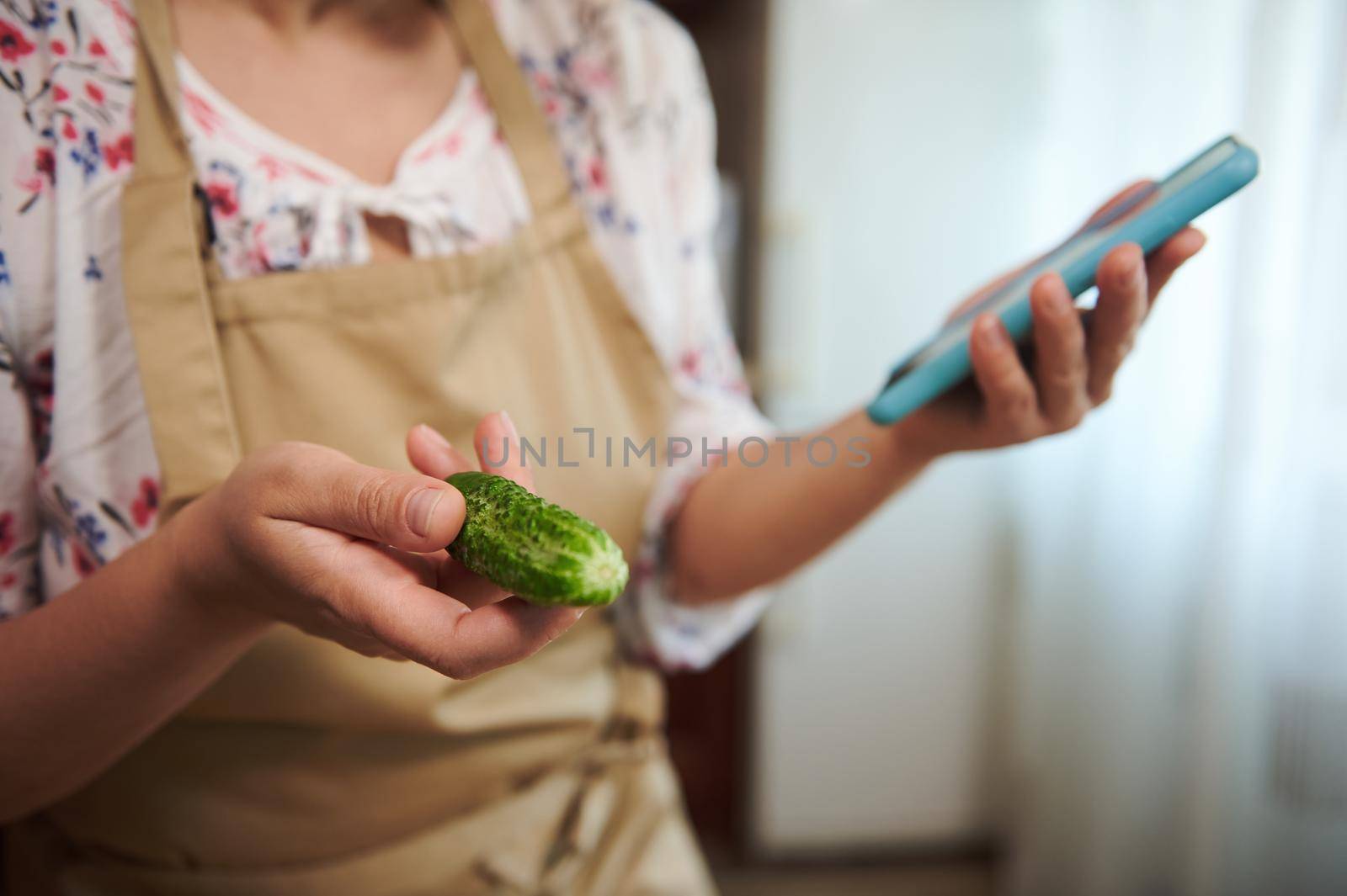 Selective focus. Fresh organic cucumber in the hand of a housewife, in a beige chef's apron using mobile phone, looks through pickling recipe in the internet web sites