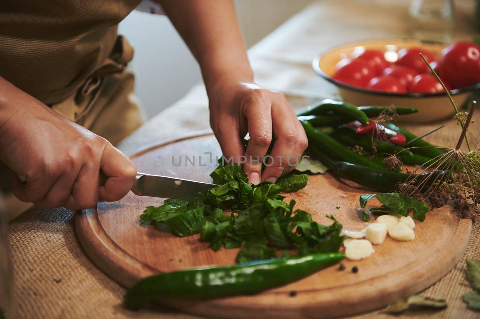 Detail: Hands of a housewife in beige chef's apron, cutting fresh fragrance culinary herbs on a wooden chopping board while preparing marinade for pickling chili peppers in the kitchen. Homemade food