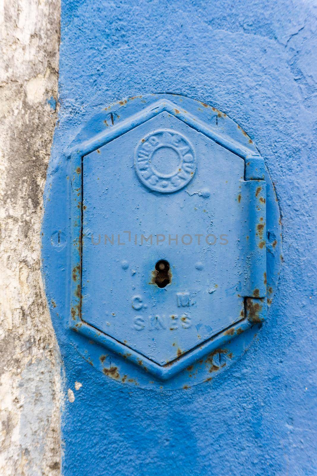 Old rusty blue painted iron lid of a valve box of the SERVICO DE AGUA PUBLIC WATER SUPPLY on the chipped white - blue painted wall of a house in the Old Town area, Portugal