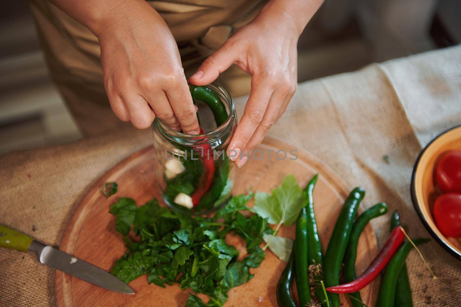 Overhead view of a housewife in a beige kitchen apron, filling sterilized glass jar with spicy chili peppers and fresh chopped fragrant ingredients when pickling seasonal vegetables in the kitchen