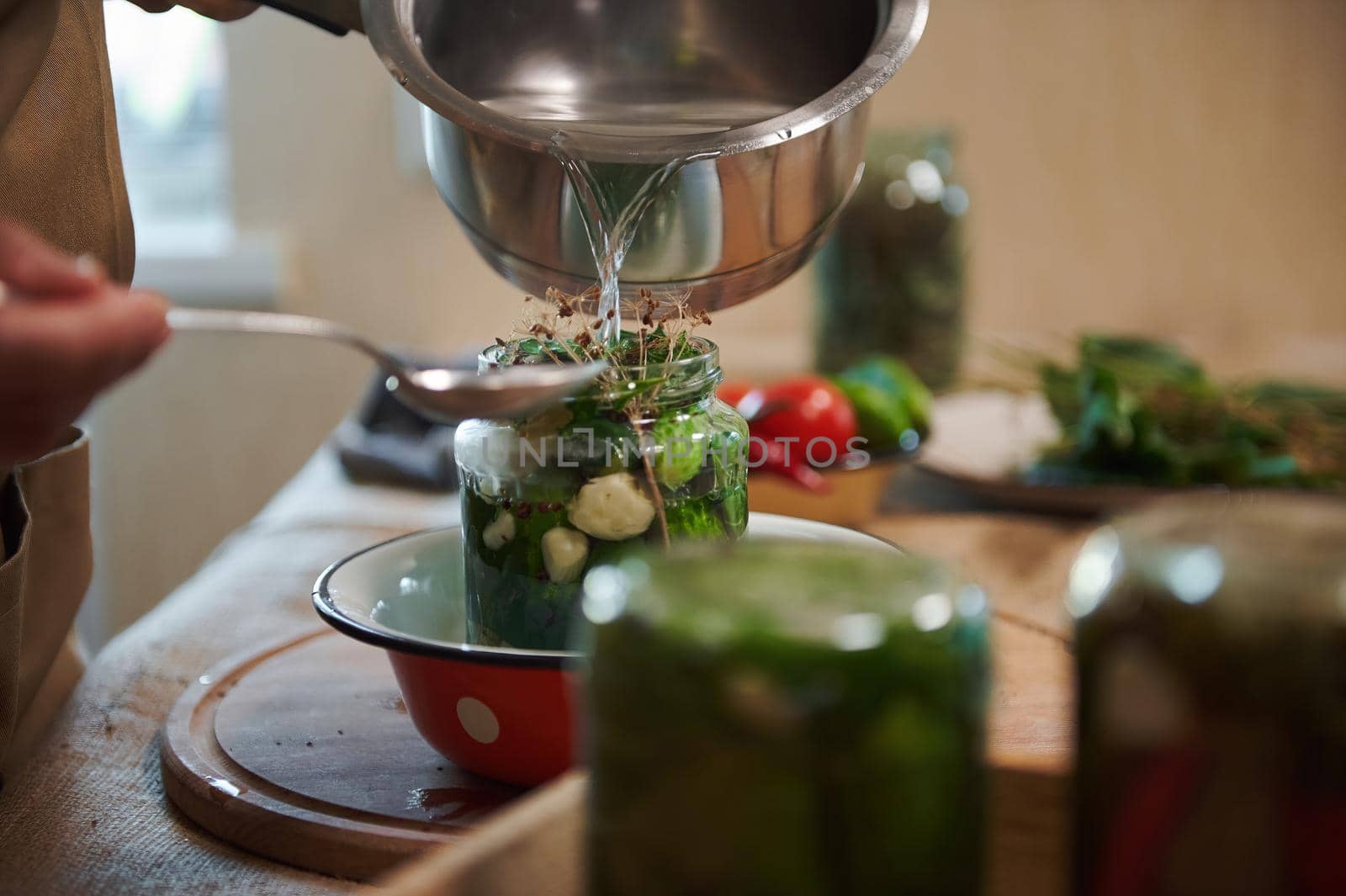 Close-up of pouring boiling marinade or brine over cucumbers, from a saucepan in jar, while pickling organic vegetable harvest at home kitchen. Jars with canned food upside down on blurred foreground