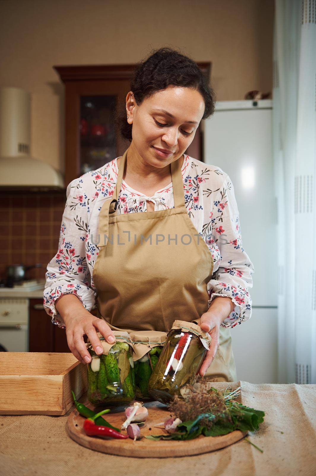 Pleasant African American woman, a housewife in beige chef's apron posing in her home kitchen with homemade pickled seasonal vegetables. Marinating. Canning. Pickling. Preservation.