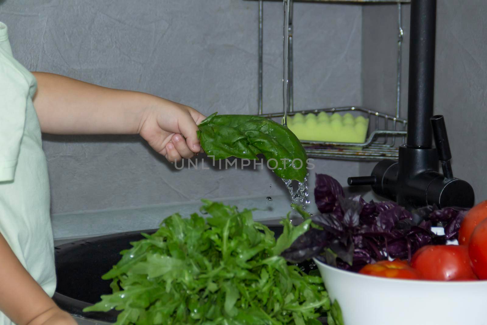 A little boy with a short haircut helps to cook in the kitchen. Washes fresh vegetables and herbs in a black sink in a gray kitchen...