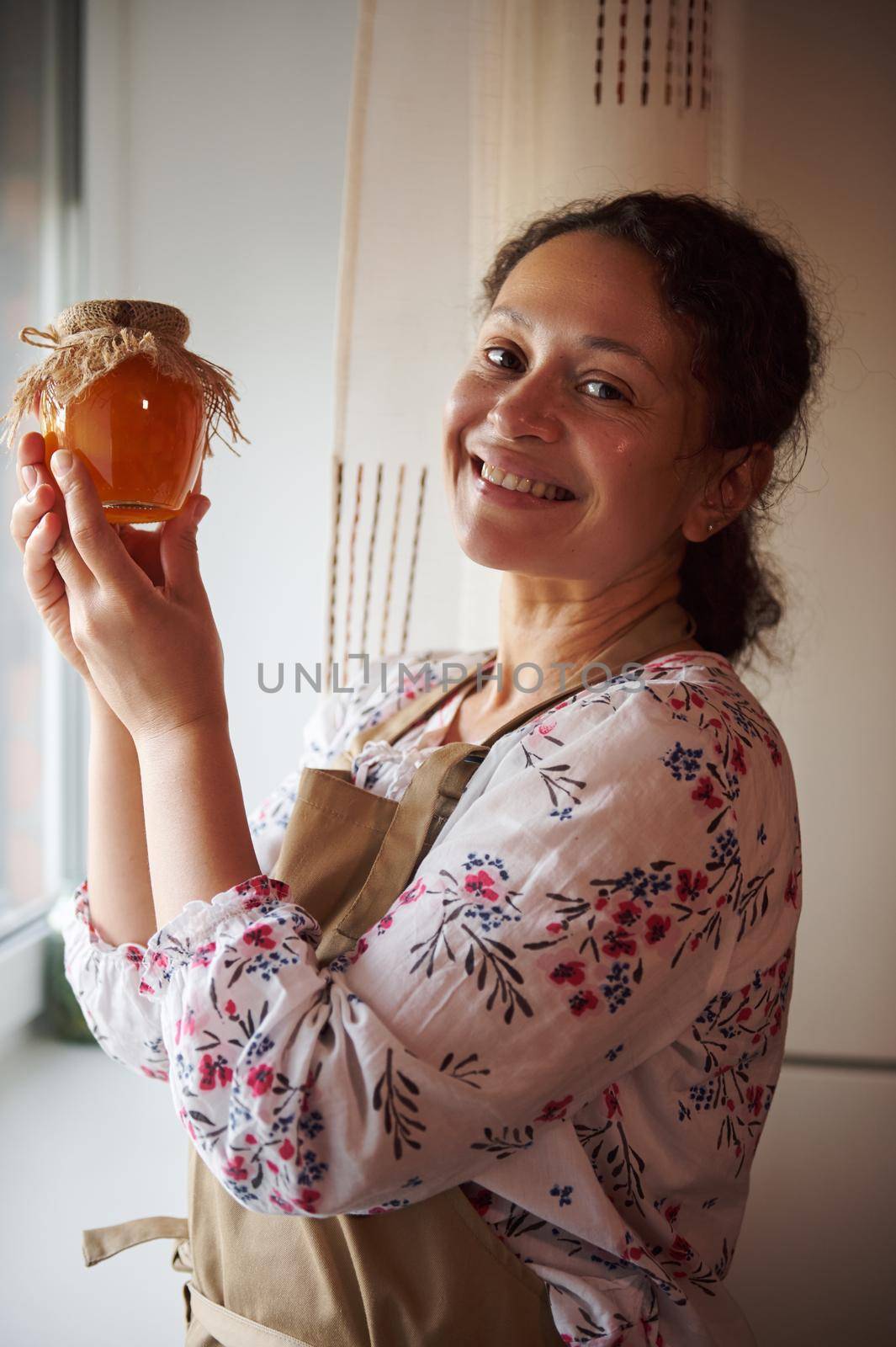 Beautiful multiethnic woman, a pleasant housewife, in a beige apron, holds a jar of homemade peach jam, canned according to a traditional family recipe, and smiles cutely, looking at the camera