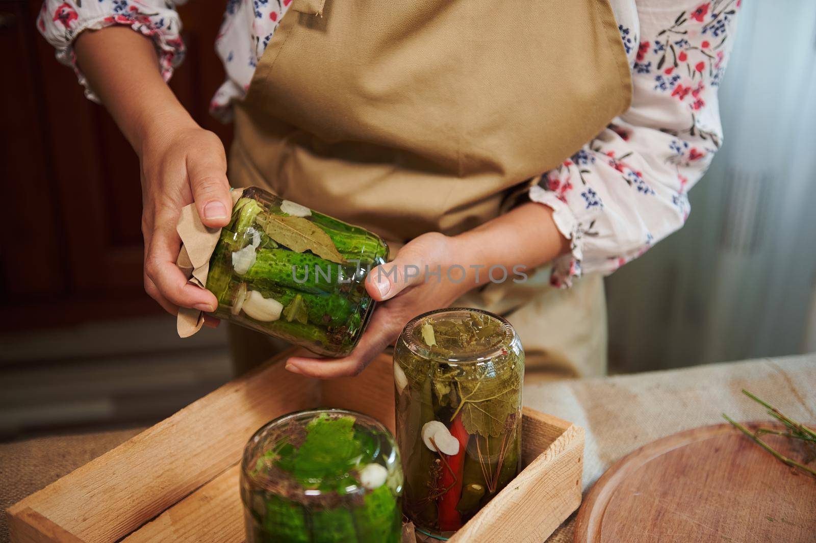 Details: female housewife's hands holding a glass caning jar of pickled cucumbers and placing it upside down on a wooden box with freshly pickled canned seasonal vegetables, in the home kitchen