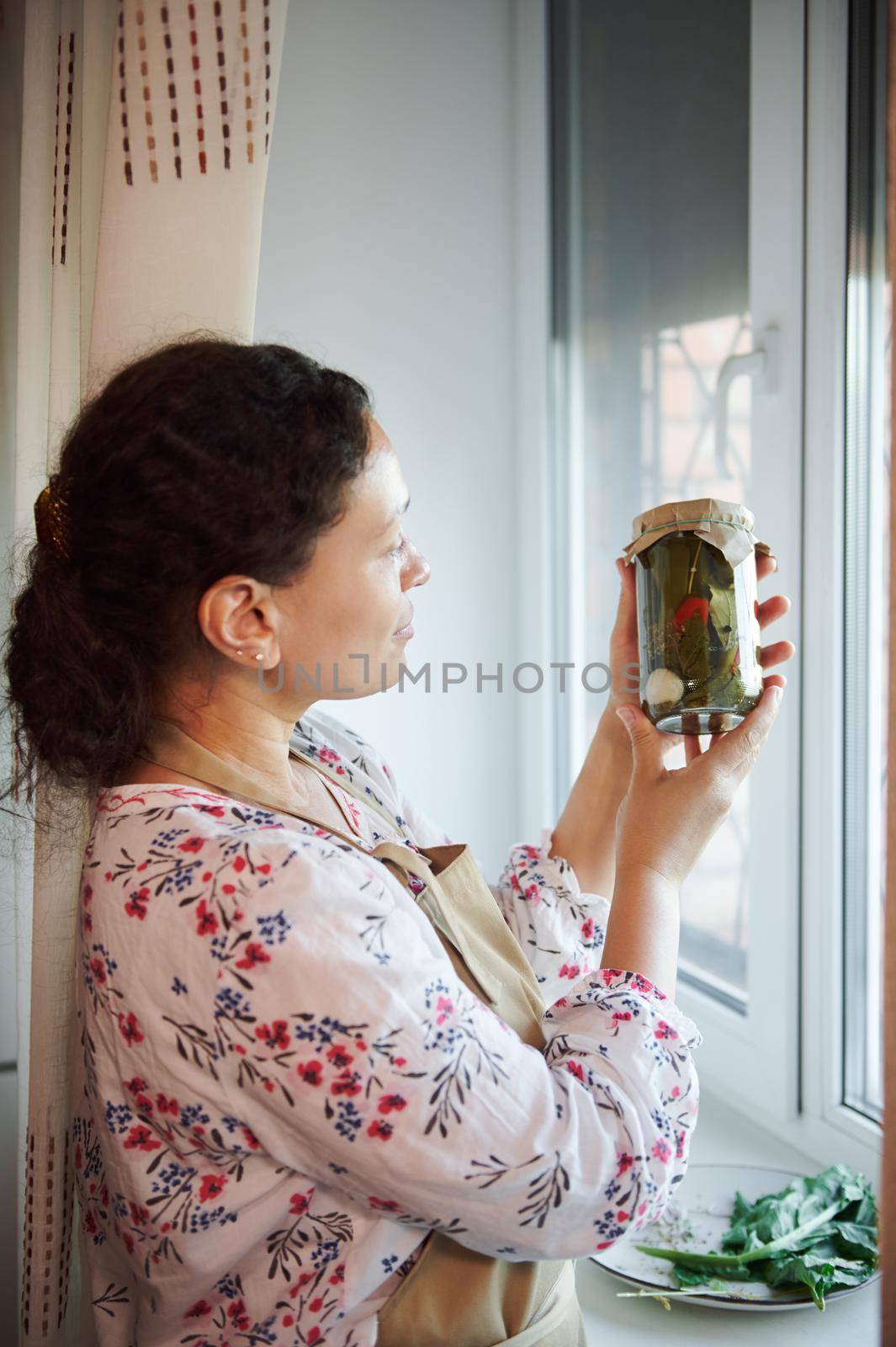 Portrait of serene mature woman, housewife in a chef's apron standing by window at home kitchen and looking at the jar with marinated chili peppers, canned according a traditional family recipe