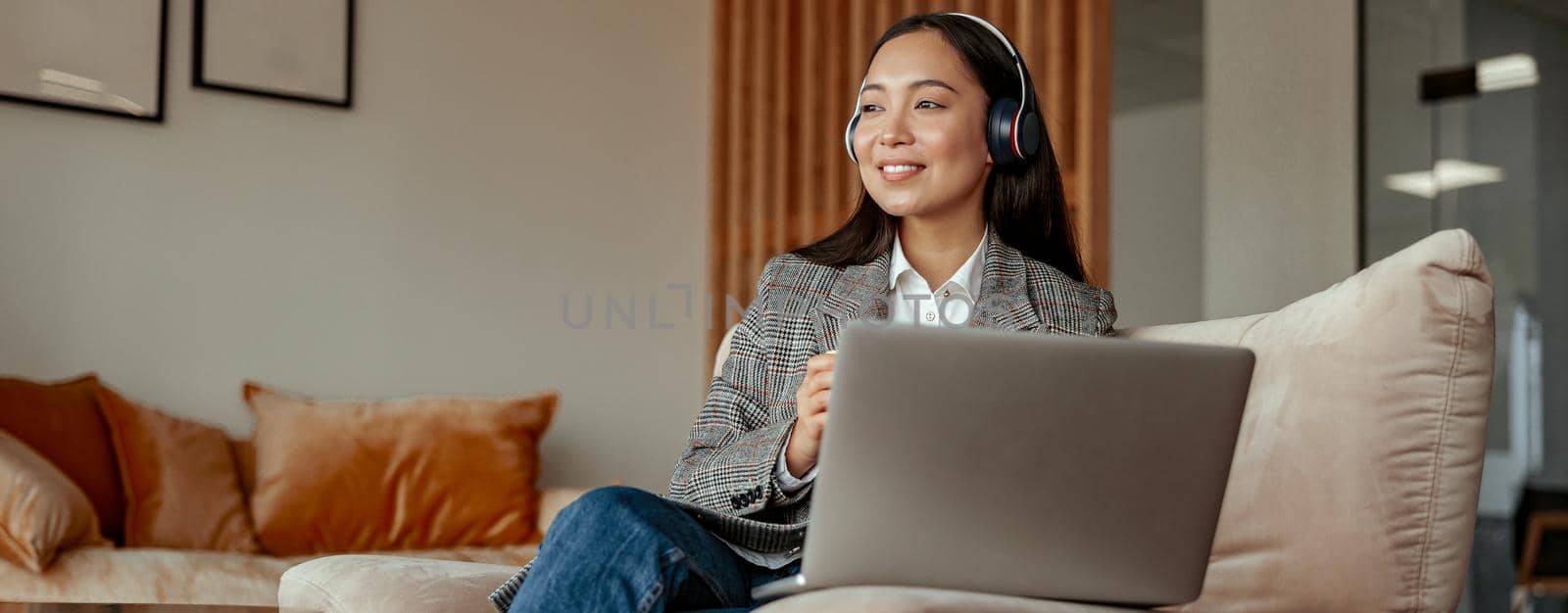 Smiling asian business woman listening music on headphones sitting with laptop during break by Yaroslav_astakhov