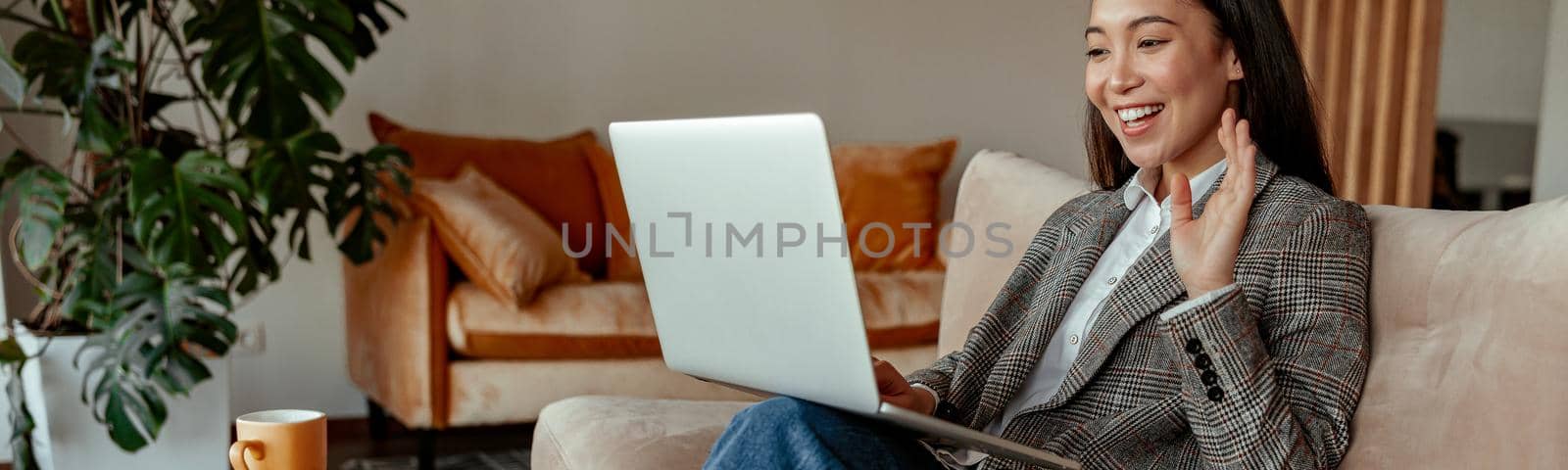 Smiling asian businesswoman making video call to client and waving greeting sit in cozy office