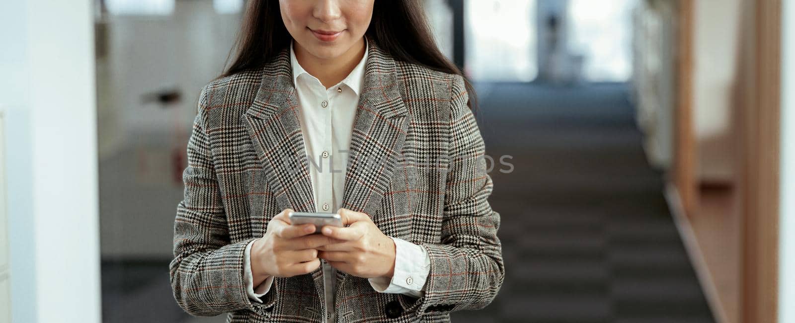 Smiling asian business woman is working correspondence on phone while standing in modern office