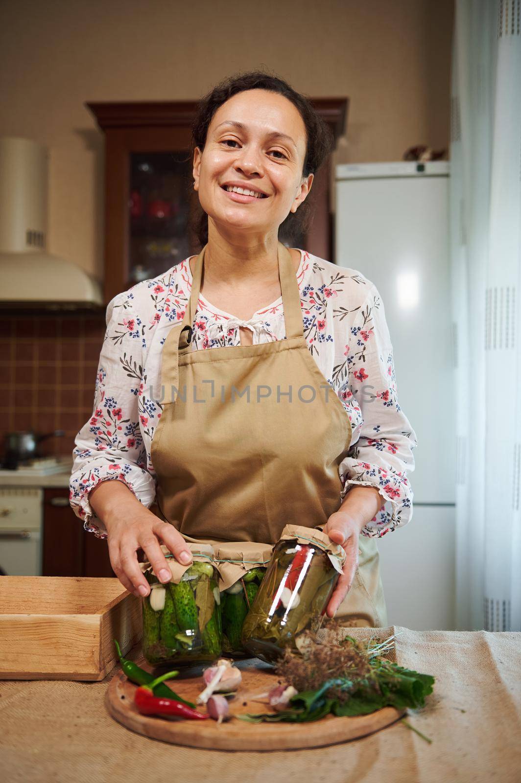 Charming multi-ethnic woman, housewife stands in the home kitchen interior and smiles looking at camera, showing jars with homemade pickled cucumbers and chili peppers. Seasonal canning of vegetables